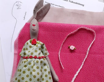 May include: A green and white polka dot dress with a red flower and a red ribbon. The dress is on a gray bunny with long ears. The bunny is on a pink fabric with white polka dots. There is a white lace trim and a pink flower on the fabric.