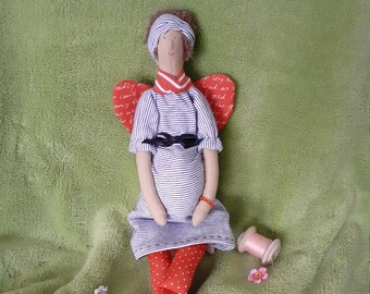 May include: A handmade doll with red wings, a striped shirt, red and white polka dot pants, and black shoes. The doll is sitting on a green fuzzy surface with spools of thread.