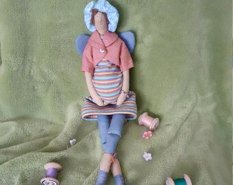 May include: A handmade fabric doll with blue wings, a pink and white striped dress, and a blue hat. The doll is sitting on a green fuzzy surface with spools of thread.