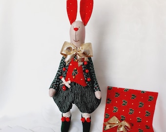 May include: A red and green fabric bunny doll wearing a gold bow tie and a red vest with gold buttons. The doll is surrounded by fabric scraps in various colors and patterns, including red, green, and white.