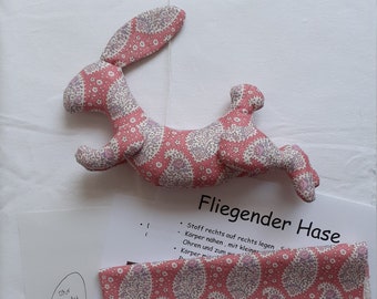May include: A pink fabric bunny with a floral pattern, lying on a white surface. The bunny is labeled "Fliegender Hase" which translates to "Flying Bunny".