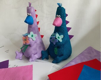 May include: Two purple and blue fabric dinosaurs holding bouquets of purple flowers. The dinosaurs are sitting on a white surface with fabric scraps in the background.