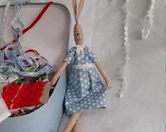Könnte beinhalten: Eine Holz-Hasenpuppe, die ein blaues Kleid mit weißen Punkten trägt. Die Puppe hat eine blaue Nase und braune Augen. Die Puppe sitzt auf einer weißen Oberfläche neben einer blauen Box.