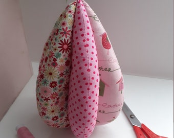 May include: A pink and white fabric Easter egg with a floral pattern, polka dots, and the word "Easter" written on it. The egg is surrounded by pink thread spools and red scissors.