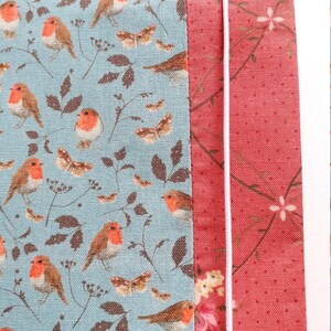 K&ouml;nnte beinhalten: Nahaufnahme eines Stoffes mit einem Muster aus Rotkehlchen und Bl&auml;ttern auf hellblauem Hintergrund. Der Stoff ist an einen roten Stoff mit einem Blumenmuster gen&auml;ht. Eine wei&szlig;e Schnur ist entlang der Kante der Stoffe gen&auml;ht.