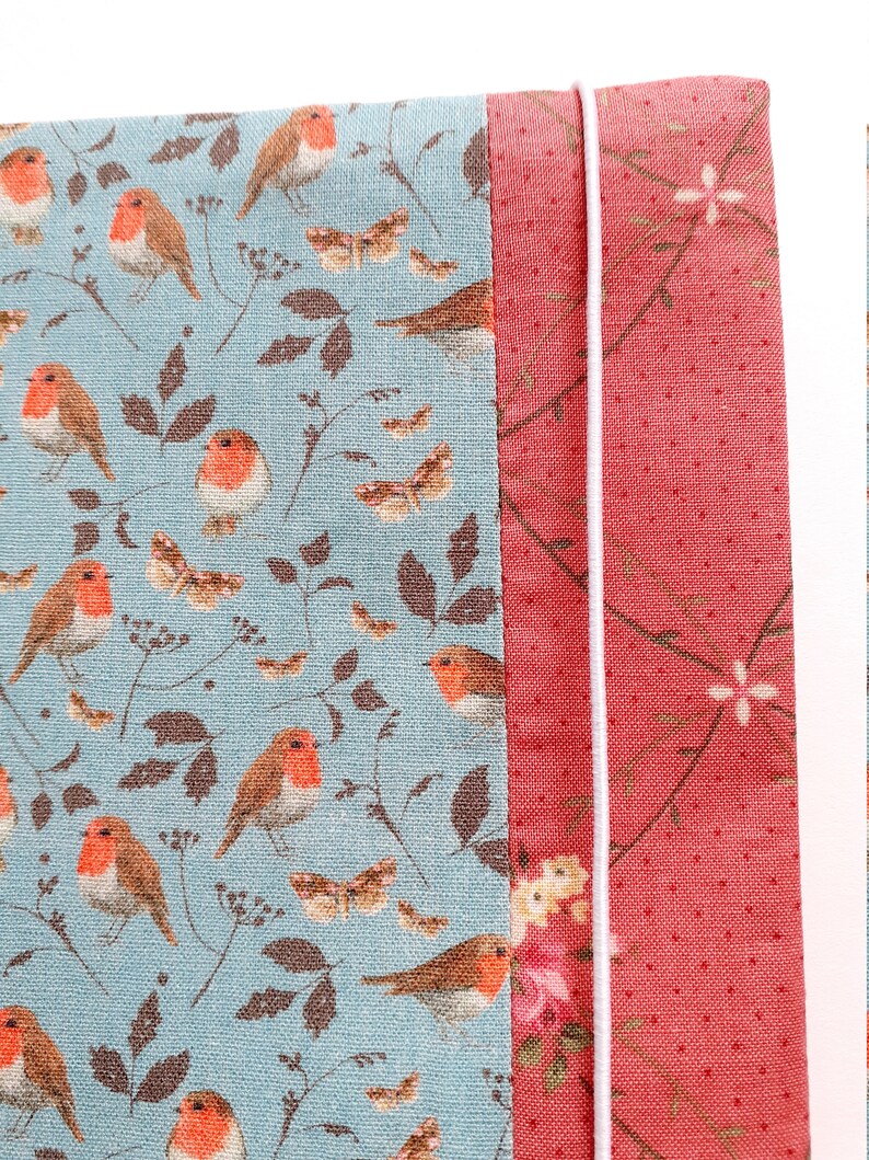 K&ouml;nnte beinhalten: Nahaufnahme eines Stoffes mit einem Muster aus Rotkehlchen und Bl&auml;ttern auf hellblauem Hintergrund. Der Stoff ist an einen roten Stoff mit einem Blumenmuster gen&auml;ht. Eine wei&szlig;e Schnur ist entlang der Kante der Stoffe gen&auml;ht.