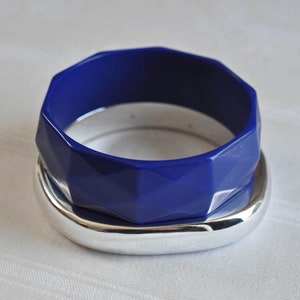 May include: A blue plastic bangle bracelet with a geometric design, stacked on top of a silver bangle bracelet.