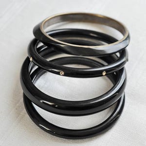 May include: A stack of four smooth, glossy black bangle bracelets. The top bracelet has a metallic inner rim, while the second bracelet from the top features small, evenly spaced clear stones embedded along its outer surface.