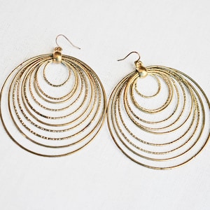May include: A pair of gold-toned dangle earrings featuring multiple concentric circular hoops. Each earring is composed of several thin, textured rings of varying sizes that are connected at the top by a small metal bead and a curved ear wire hook.