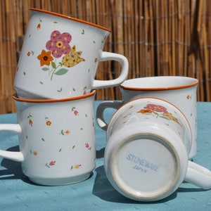 May include: Four white ceramic mugs with floral designs. The mugs have a brown rim and are stacked on top of each other. The mug on the bottom has the words "Stoneware Japan" printed on the bottom.