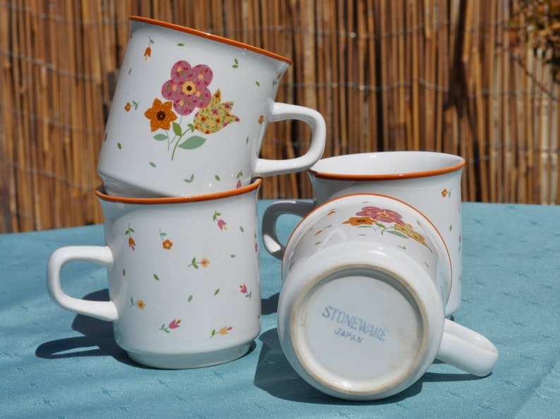 May include: Four white ceramic mugs with floral designs. The mugs have a brown rim and are stacked on top of each other. The mug on the bottom has the words "Stoneware Japan" printed on the bottom.