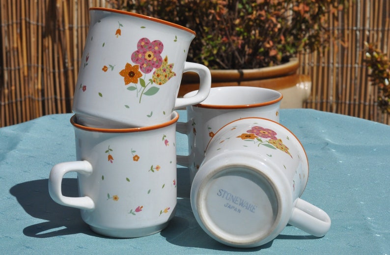 May include: Four white ceramic mugs with floral designs. The mugs are stacked on top of each other. The bottom mug has the words "Stoneware Japan" printed on the bottom.