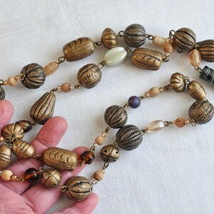 May include: A long necklace made of brown, gold, and white beads. The beads are various shapes and sizes, including round, oval, and rectangular. Some of the beads have a textured surface.