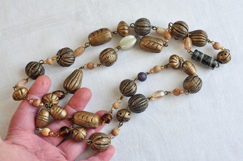 May include: A long necklace made of brown, gold, and white beads. The beads are various shapes and sizes, including round, oval, and rectangular. Some of the beads have a textured surface.