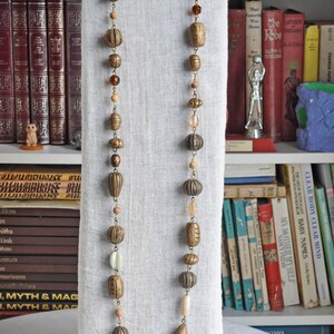 May include: A long necklace with brown, gold, and white beads. The beads are various shapes and sizes, including round, oval, and teardrop. The necklace is displayed on a white fabric stand.