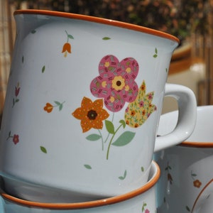 May include: Set of three white ceramic mugs with orange rims. Each mug features a different floral design with pink, yellow, and orange flowers.