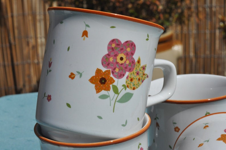 May include: Set of three white ceramic mugs with orange rims. Each mug features a different floral design with pink, yellow, and orange flowers.