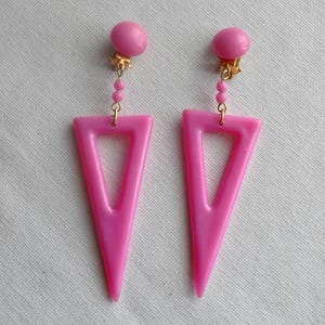 May include: A pair of pink triangle shaped dangle earrings with gold tone accents. The earrings have a clip on closure.