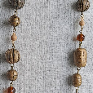 May include: A long necklace with brown and gold beads, including round and rectangular shapes, on a white fabric background.