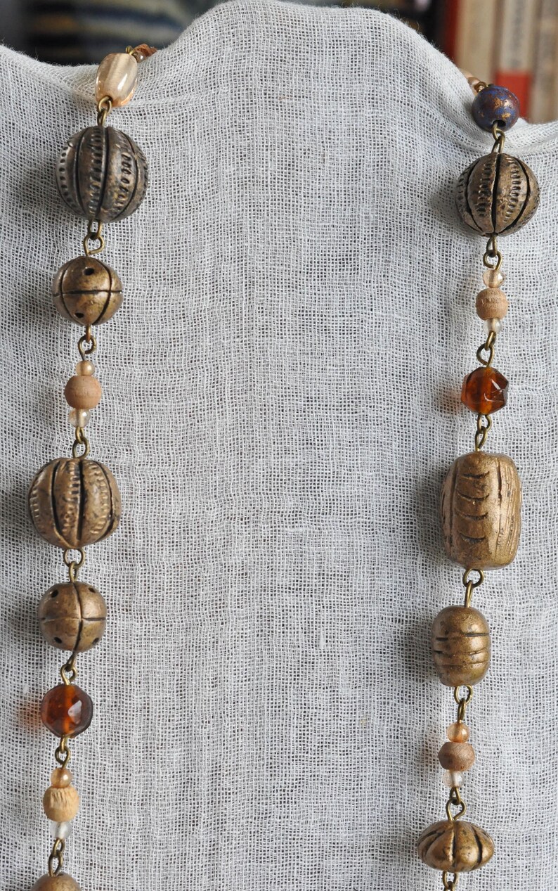 May include: A long necklace with brown and gold beads, including round and rectangular shapes, on a white fabric background.