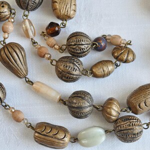 May include: A long, multi-strand necklace with brown, beige, and white beads. The beads are made of various materials, including metal, wood, and glass. The necklace is strung on a silver chain.