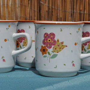 May include: Four white ceramic mugs with floral designs in shades of pink, yellow, and orange. The mugs have brown rims and handles.
