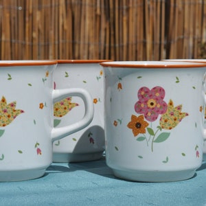 May include: Four white ceramic mugs with floral designs. Each mug features a different floral pattern with pink, yellow, and orange flowers. The mugs have a brown rim and handle.