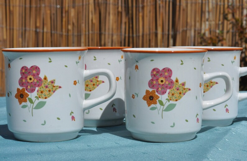 May include: Four white ceramic mugs with floral designs. Each mug features a different floral pattern with pink, yellow, and orange flowers. The mugs have a brown rim and handle.