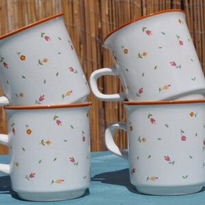 May include: Four white ceramic mugs with a floral pattern in shades of pink, orange, and yellow. The mugs are stacked on top of each other.