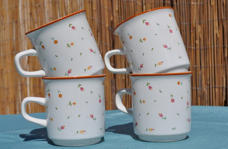 May include: Four white ceramic mugs with a floral pattern in shades of pink, orange, and yellow. The mugs are stacked on top of each other.