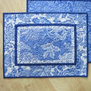 May include: Two blue and white quilted placemats with a floral pattern. The placemats have a rectangular shape with a dark blue border. The floral design features leaves and flowers in shades of blue and white.