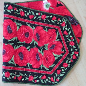 May include: A quilted table runner featuring a vibrant poppy design. The runner has a black border with red poppies and green leaves. The background is red with a poppy pattern. The edges are trimmed with green.