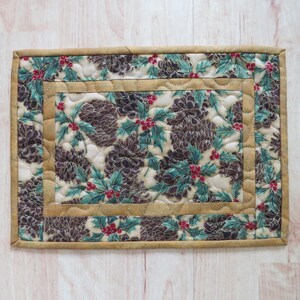 May include: A rectangular quilted placemat with a festive holiday design. The fabric features a pattern of brown pinecones, green holly leaves, and red berries on a cream background. The edges are trimmed with a tan border.
