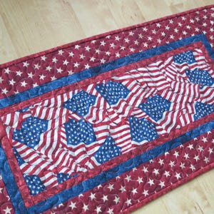 May include: A rectangular quilted table runner featuring a patriotic design. The center showcases a pattern of American flags in red, white, and blue. The border is a deep red with white stars, and a blue border frames the flag design.
