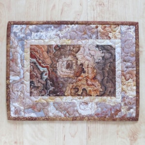May include: A rectangular, quilted fabric piece with a marbled agate design. The central area features a detailed pattern in shades of brown, red, and cream, framed by a white border. The outer border is a darker brown, and the piece is set on a light wood surface.