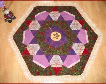 May include: A hexagonal Christmas tree skirt with a white lace trim. The skirt is made of various fabrics in shades of purple, green, and red. A gold starfish is in the center of the skirt.