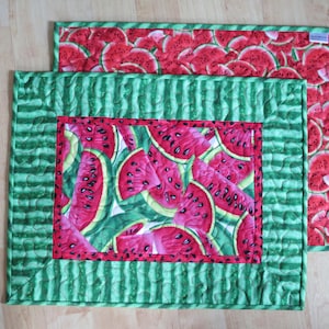 Puede incluir: Dos manteles individuales acolchados de color verde y rojo con un patr&oacute;n de sand&iacute;a. Los manteles individuales tienen un borde de rayas verdes y blancas y un borde interior rojo. El patr&oacute;n de sand&iacute;a est&aacute; en el centro de los manteles individuales.