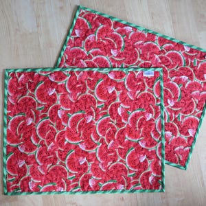 Puede incluir: Dos manteles individuales de tela roja y verde con estampado de sand&iacute;a y borde verde. Los manteles individuales son de tela y tienen una textura acolchada.