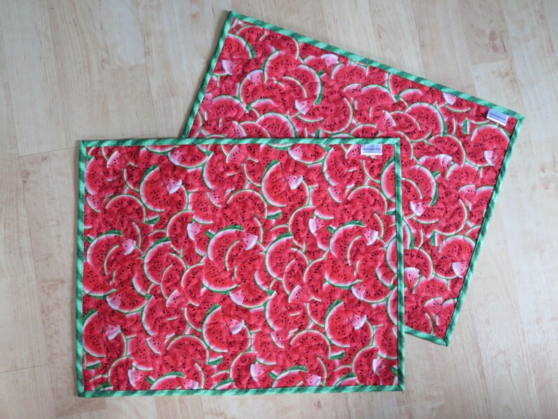 Puede incluir: Dos manteles individuales de tela roja y verde con estampado de sand&iacute;a y borde verde. Los manteles individuales son de tela y tienen una textura acolchada.