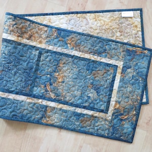 May include: A rectangular quilted table runner featuring a marbled pattern in shades of deep blue, tan, and cream. The fabric has an all-over stippled quilting design with a dark blue border and a thin cream accent strip framing the center.