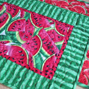 Puede incluir: Un mantel individual de tela acolchada verde y rojo con un patr&oacute;n de sand&iacute;a. El mantel individual tiene forma cuadrada y un borde rojo.