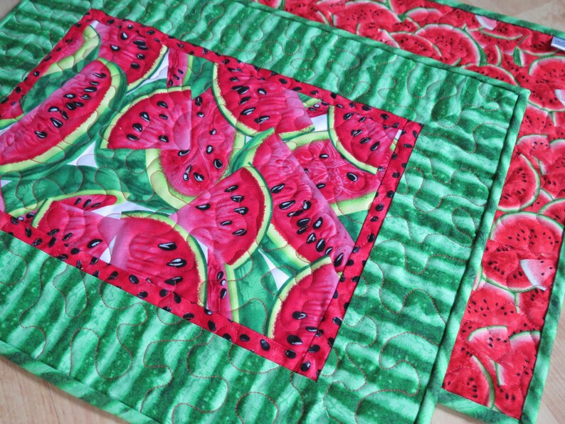 Puede incluir: Un mantel individual de tela acolchada verde y rojo con un patr&oacute;n de sand&iacute;a. El mantel individual tiene forma cuadrada y un borde rojo.