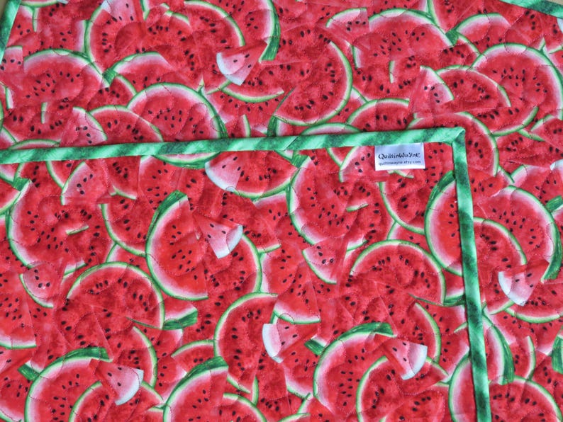 Puede incluir: Una tela acolchada verde y blanca con estampado de sand&iacute;a. La tela presenta un patr&oacute;n repetitivo de rodajas de sand&iacute;a rojas con semillas negras.
