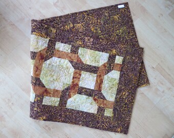 May include: A brown and cream quilted table runner with a geometric pattern. The runner has a brown border with a leaf pattern.