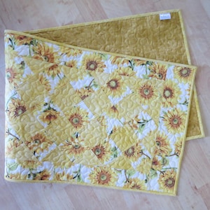 May include: A yellow table runner with a quilted pattern featuring a sunflower design. The runner has a yellow border and a solid yellow back.