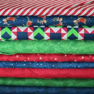 May include: A stack of eight different fabric prints in a variety of Christmas themes. The fabrics include red and white stripes, blue with reindeer, green with white dots, red with white stars, blue with white dots, red, green, and blue.
