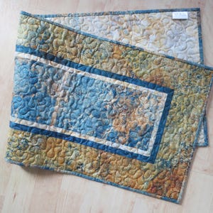 May include: A rectangular quilted table runner featuring a mottled pattern in shades of blue, gold, and tan. The fabric has a stippled texture with a dark blue border framing a central rectangular panel. A small white fabric label is attached at the corner.
