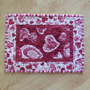 May include: A quilted placemat featuring a heart pattern in shades of pink and white. The center has large heart shapes, while the border has smaller hearts and striped accents. The placemat is rectangular.