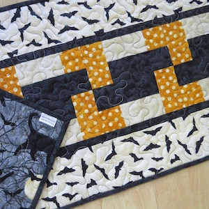 May include: A black and white fabric table runner with a black and orange geometric pattern. The runner is decorated with a repeating pattern of bats. The fabric has a quilted texture. The runner is labeled "Quiltin'Jodi".