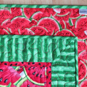 Puede incluir: Una tela acolchada verde y roja con un patr&oacute;n de sand&iacute;a. La tela tiene un borde verde y una etiqueta que dice "QuiltsbyJeni QuiltsbyJeni.com".